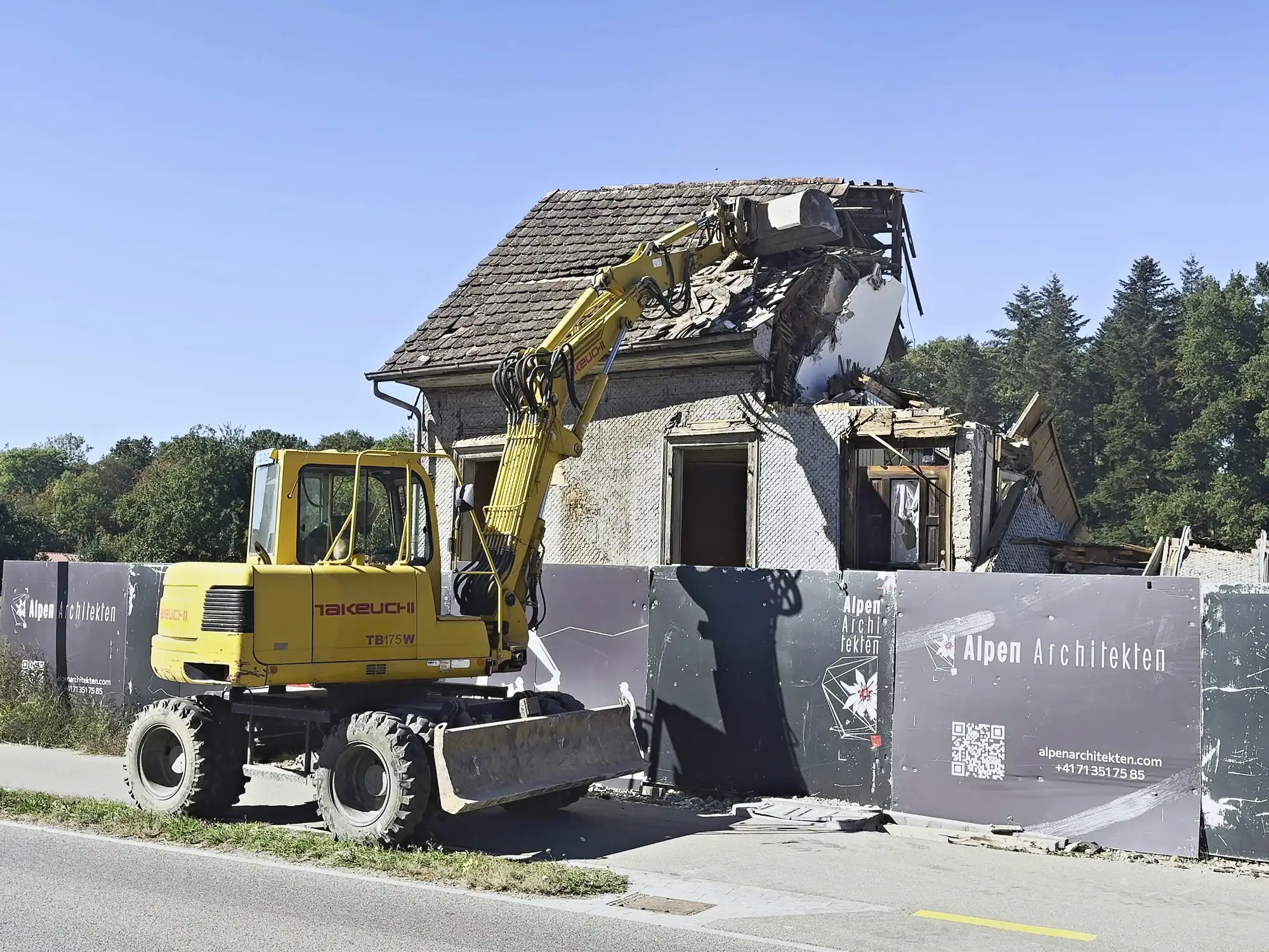Bagger vor dem halb abgebrochenen Altbau an der Strasse, Bauzaun mit dem Schild des Architekturbüros