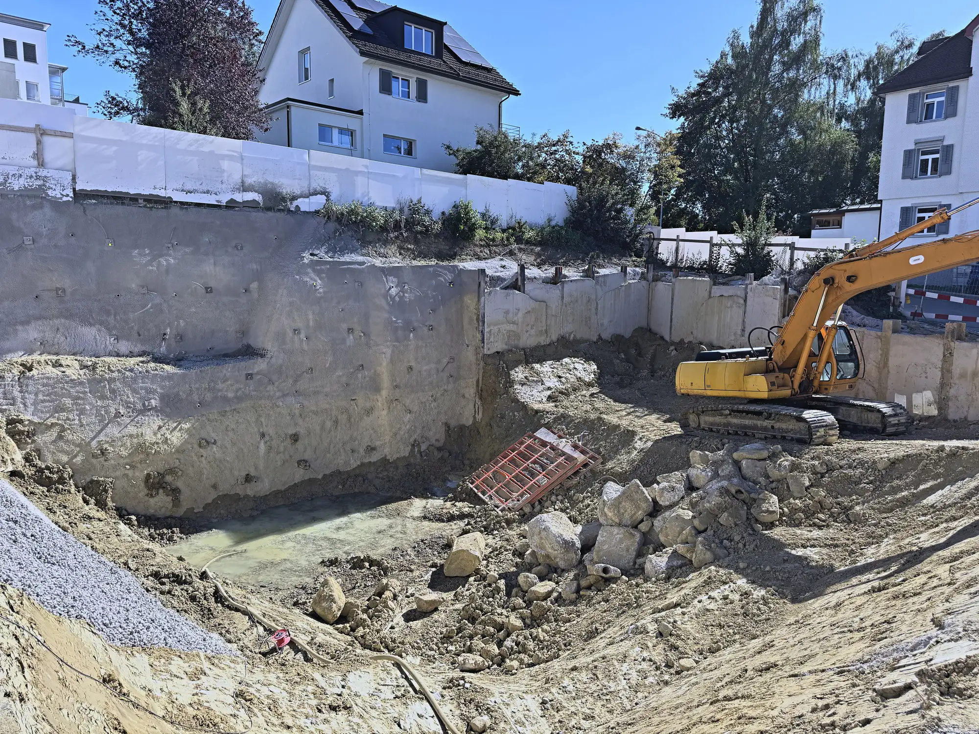 Aushubsohle mit Wasser, gesicherte Baugrubenwand und Bagger, dahinter die Nachbarhäuser