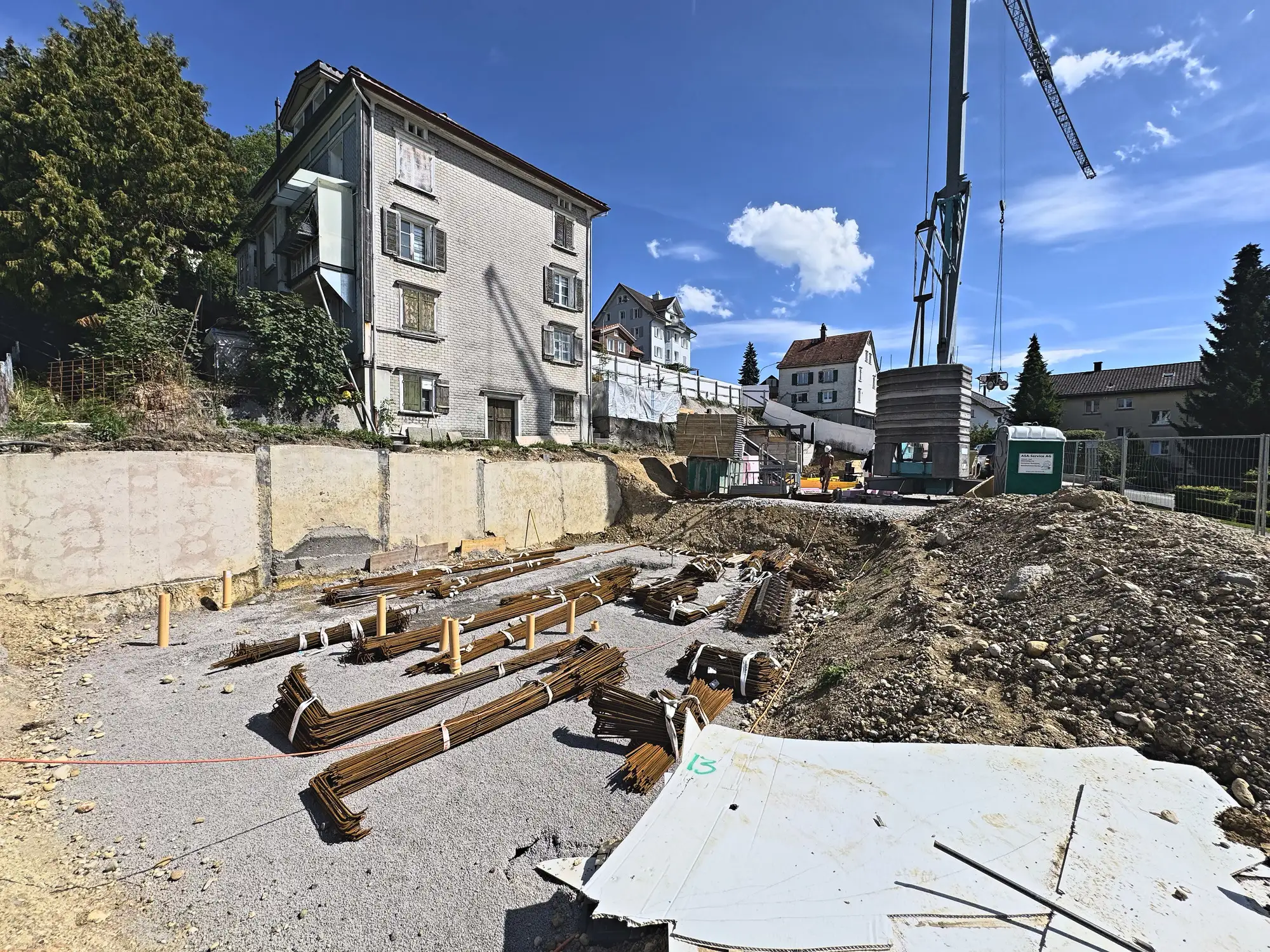 Gebündelter Bewehrungsstahl auf dem Planum, dahinter Stützmauer und Bestandsbau